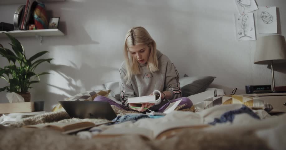 A young woman studies using a laptop and a textbook while sitting on her bed during the day
