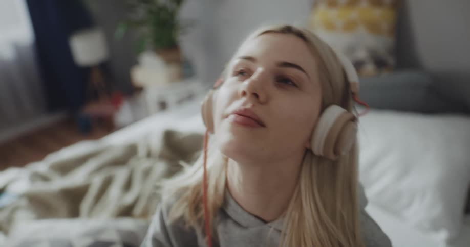 Blonde woman with gray hoodie and purple pants listens to music with headphones on bed in room