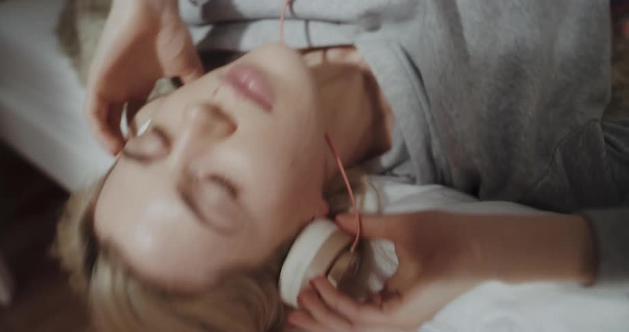 A young woman resting in a bed places a pair of headphones over her ears