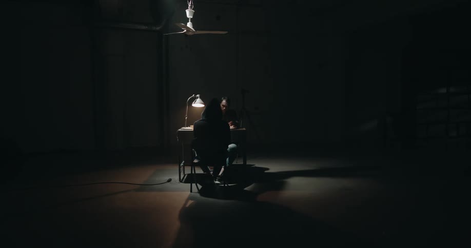 A ceiling fan slowly spins over two men sitting at a desk as one stretches his arms