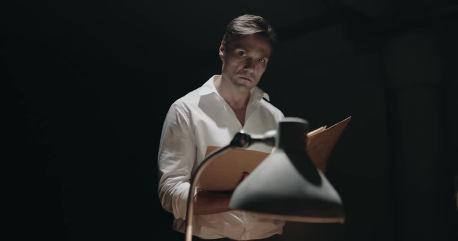 Two men sit at a desk and argue as another man reading a file walks up and stares at them