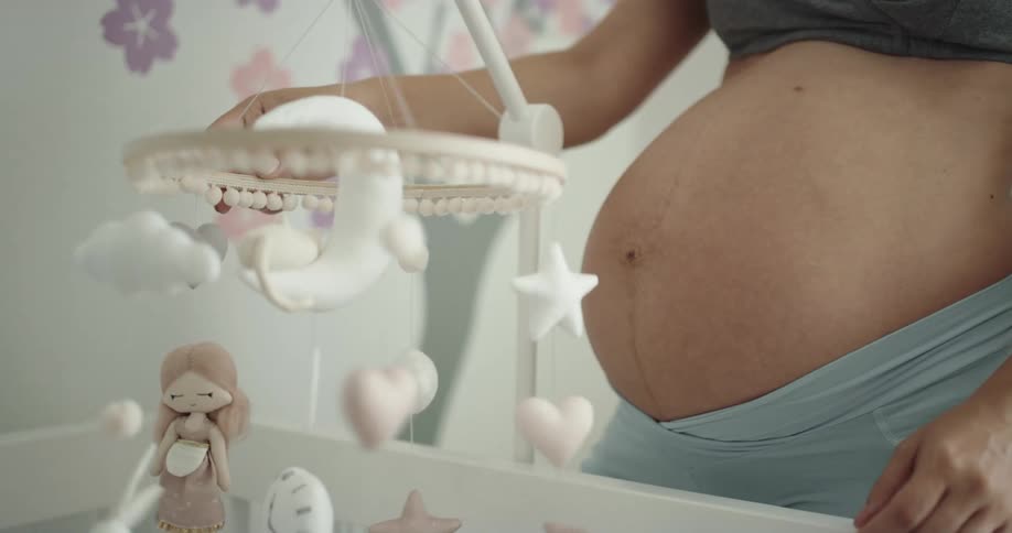 A pregnant woman examines the mobile hanging over a crib