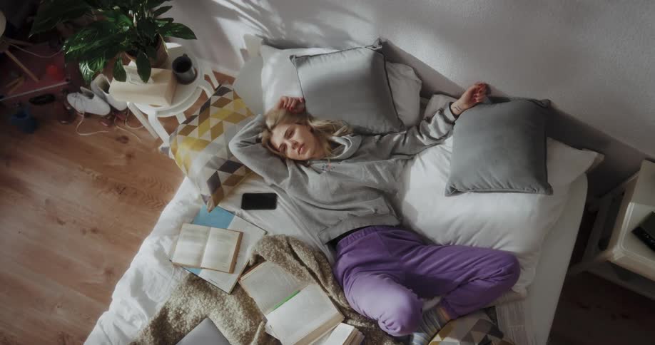 A woman rests in a messy bed covered in books while lost in thought