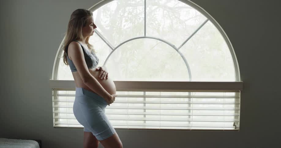 A woman is standing by a window and rubbing her pregnant belly