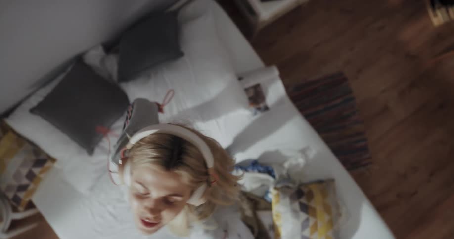 Blonde woman with gray sweater and purple pants listens to music with headphones dancing on bed