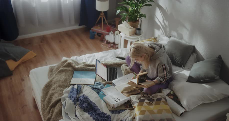 A young woman consults several books while studying in a bedroom during the day