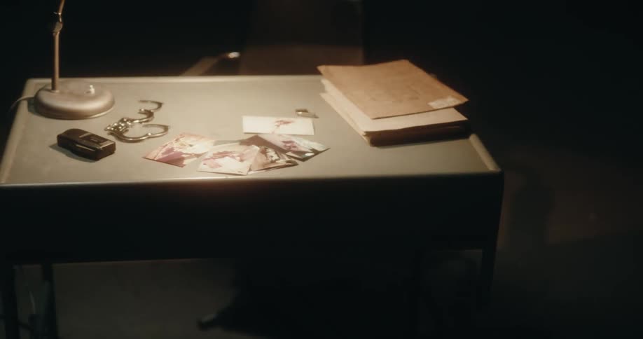 Desk lamp lights crime scene photographs and other items lying on desk in dark interrogation room