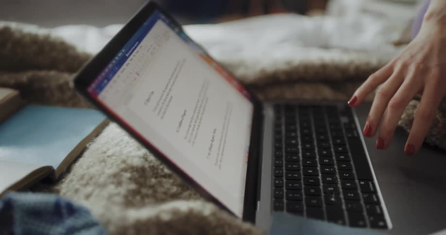 A person sits on their bed with a laptop and multiple large books