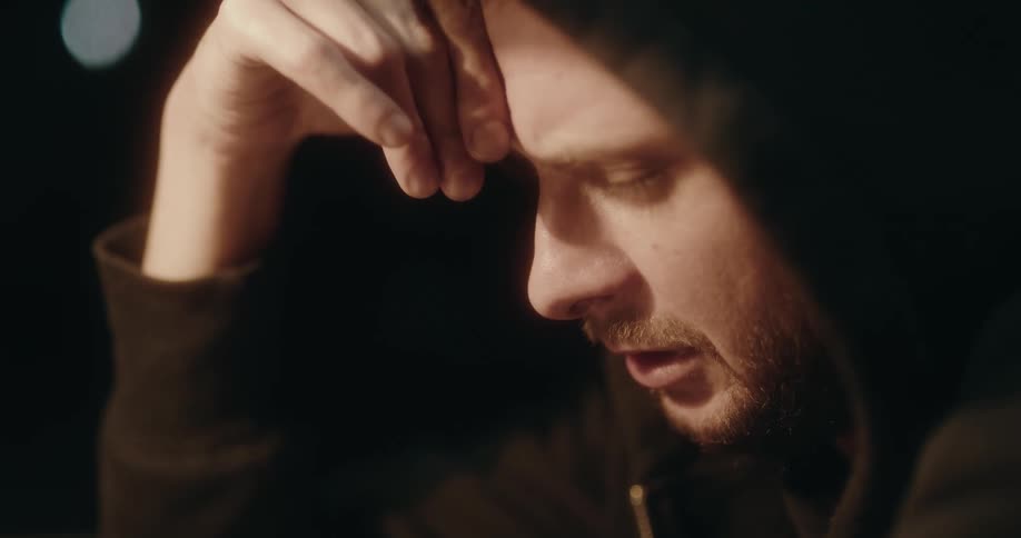 Man wearing hoodie sits in dark room lit by desk lamp and looks downcast