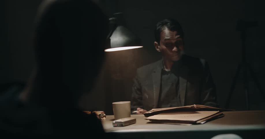 Police detective smokes and drinks while handling files and talking to a suspect in a dark interview room