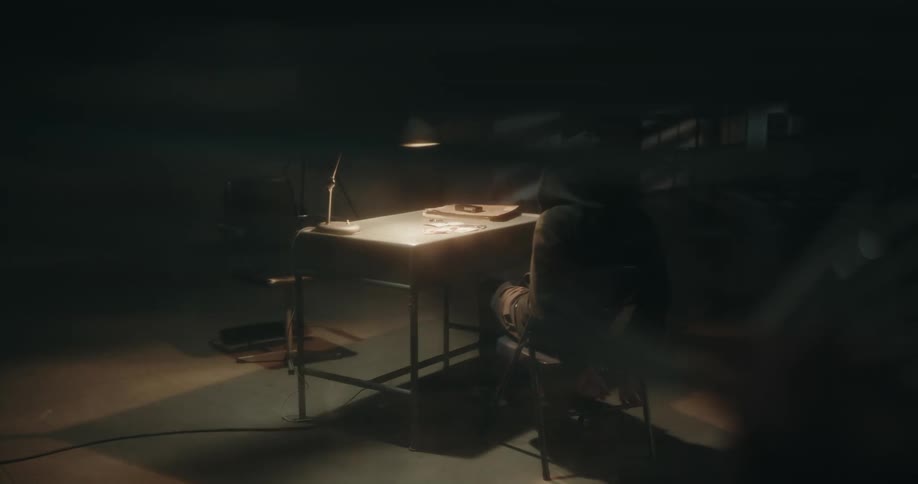 Man looks through a window into an interrogation room where a man is handcuffed to a chair