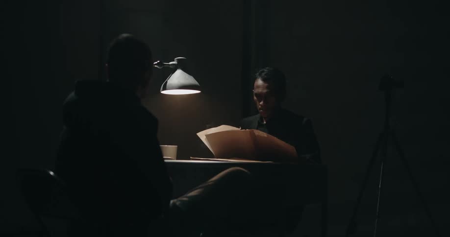 Detective reads a file while smoking in front of a suspect on the interrogation room