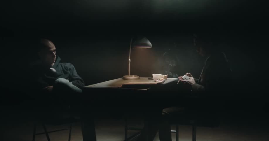 Male interviewer lights cigarette then picks up file as he sits across table in interview room from another man