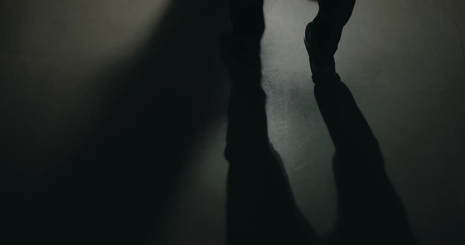 Close up of a mans legs as he walks in a building with the shadow of a fan above spinning on the floor