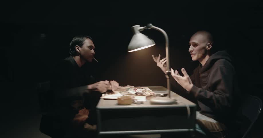Two men sit at a desk as one man smokes and shows the other evidence and they argue