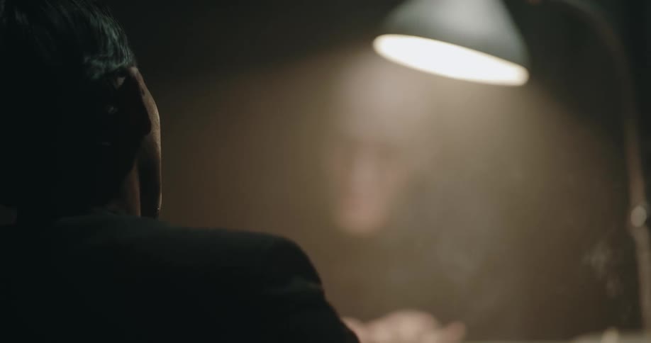 Man sits smoking in dim room across table from gesticulating suspect