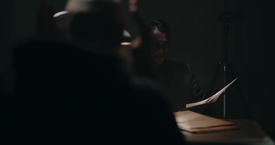 A man sits at a dark table with another man smoking a cigarette looking through papers