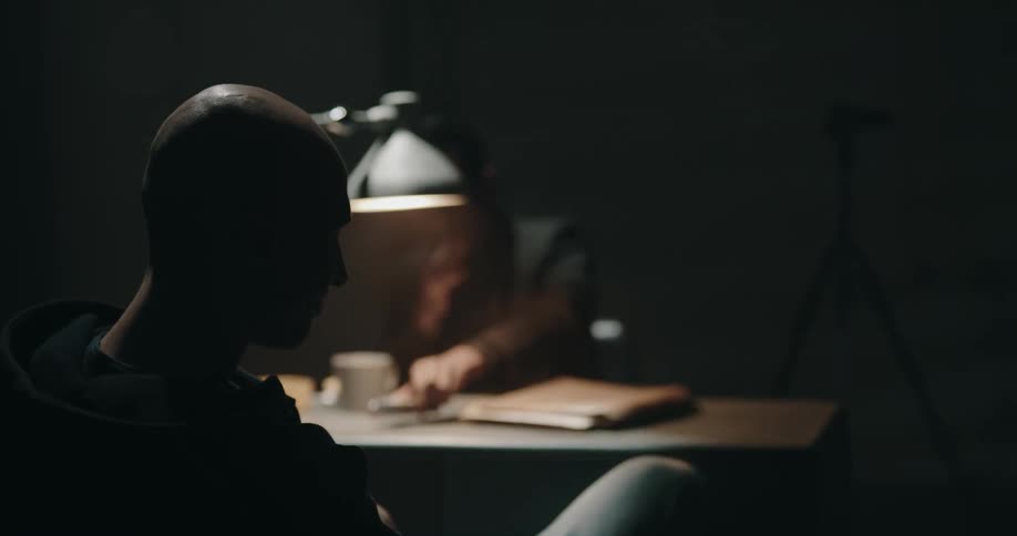 Man lights a cigarette and starts reading a file while sitting inside an interrogation room