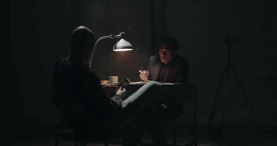 Police detective sits across from man looking through crime scene photographs in a dark interview room