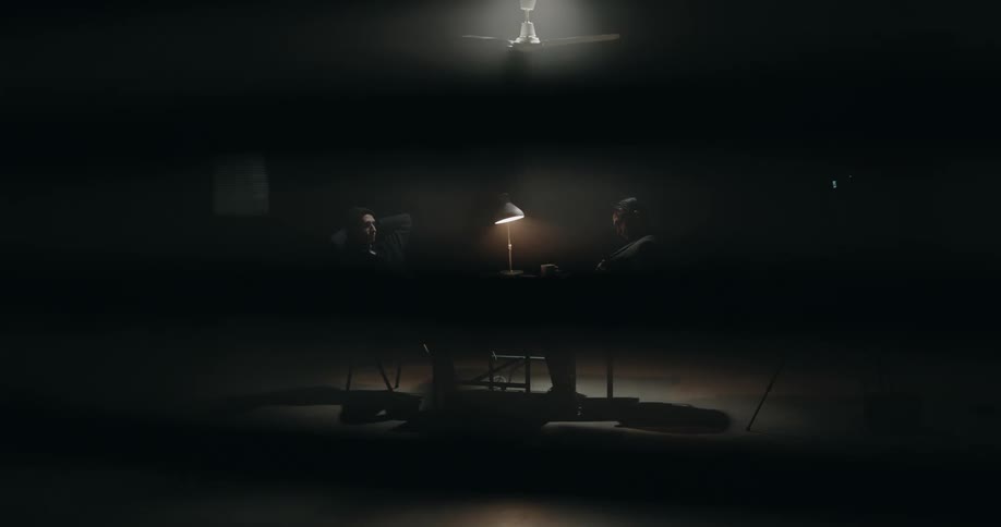 Male policeman walks into dark interrogation room to take a seat across from man at table with single desk lamp