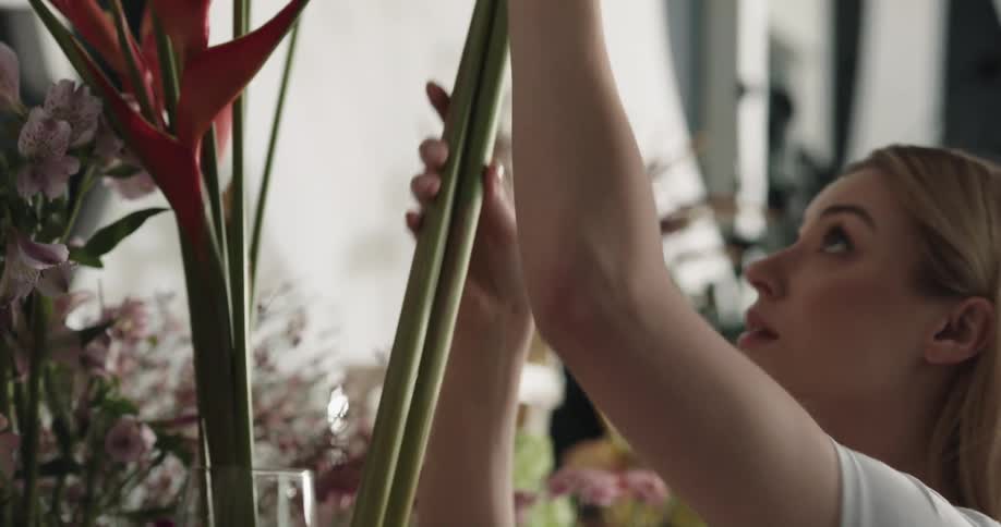 A young woman removes a large flower from a glass vase before turning and walking out of frame