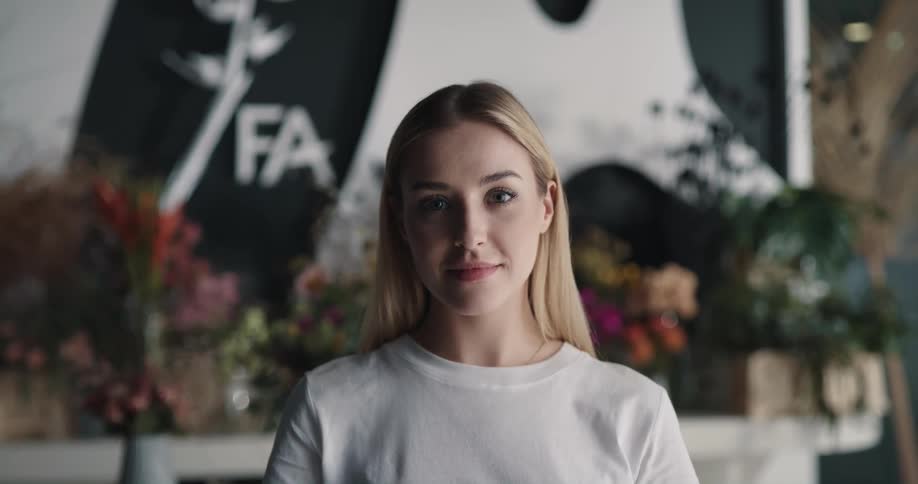 A woman standing before a shelf holding flower arrangements raises her head to smile into the camera