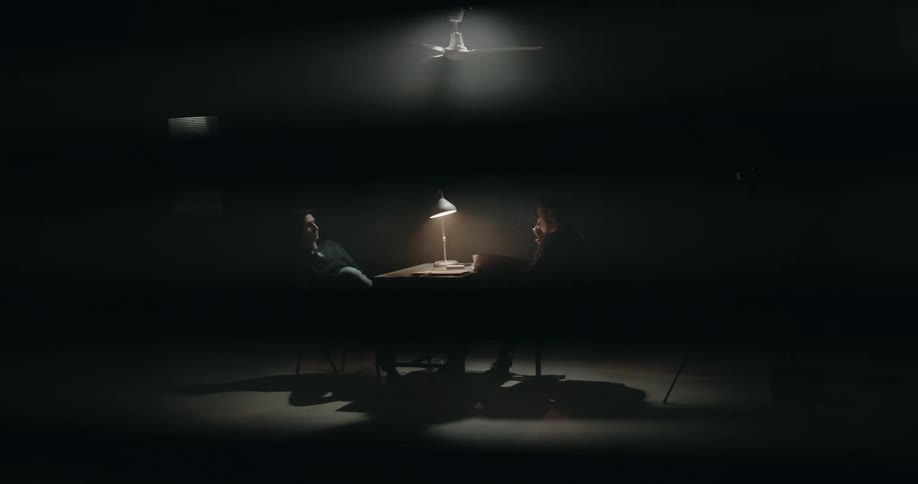 Two men sitting at a small table in a dark room with a ceiling fan above