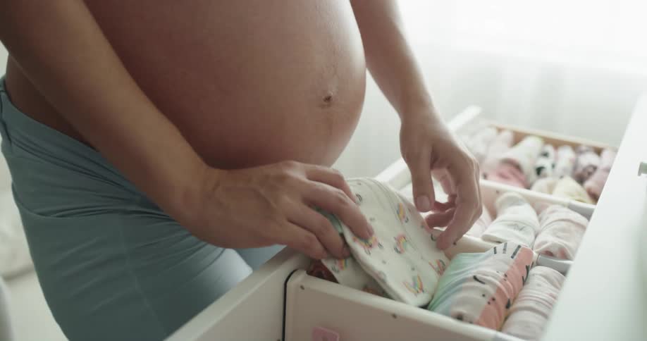 A pregnant woman opens a drawer filled with baby clothing and rearranges the contents