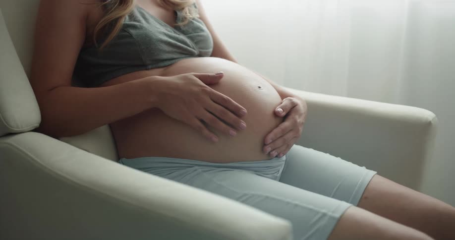 A pregnant woman rubs her stomach while sitting in a rocking chair indoors during the day