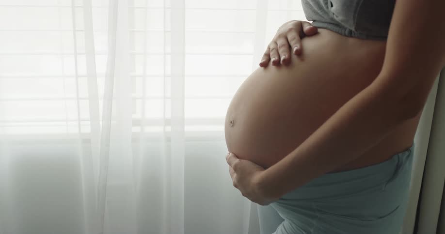 Pregnant woman with gray top and blue pants stands next to window with curtains and strokes her belly in room at home