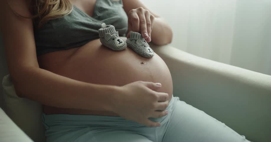 Pregnant woman with green top puts baby socks on her belly and swings in armchair