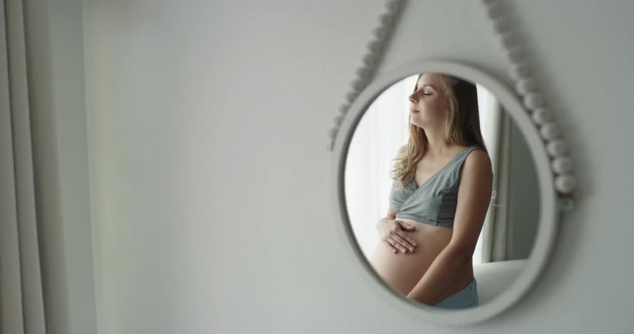 Blonde pregnant woman with gray top strokes her belly seen from small hanging mirror mirror