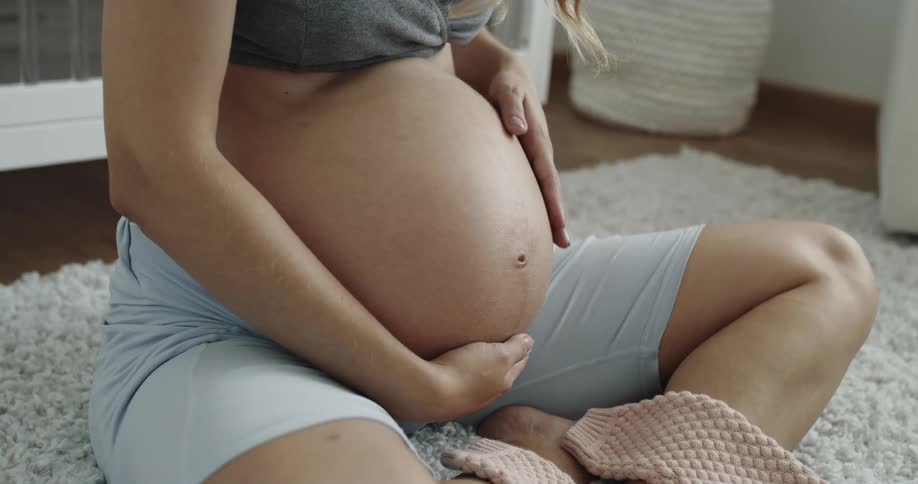 A pregnant woman rubs her belly with her hands while sitting on a rug indoors during the day