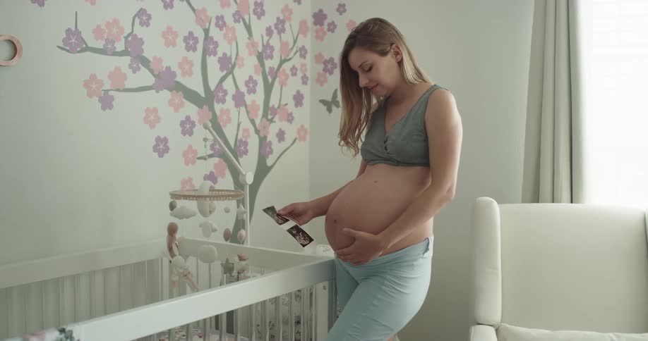 A pregnant woman rubs her belly while looking at printed sonogram images as she stands next to a crib indoors during the day