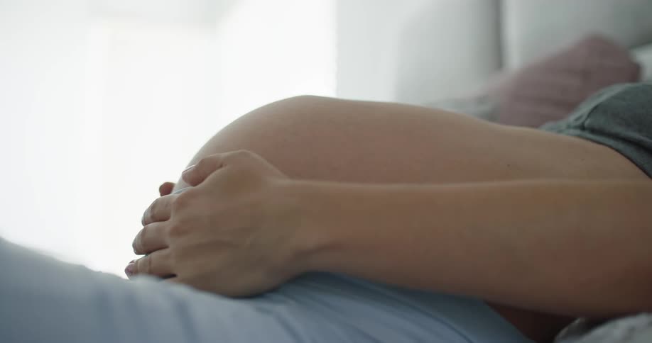 Pregnant woman with green top and blue shorts lies on bed holding her belly