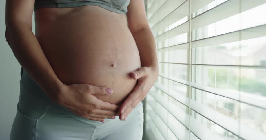 Pregnant woman standing next to a window rubbing her belly with both hands