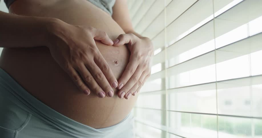 A pregnant woman rests her hands on her stomach while looking out of a window