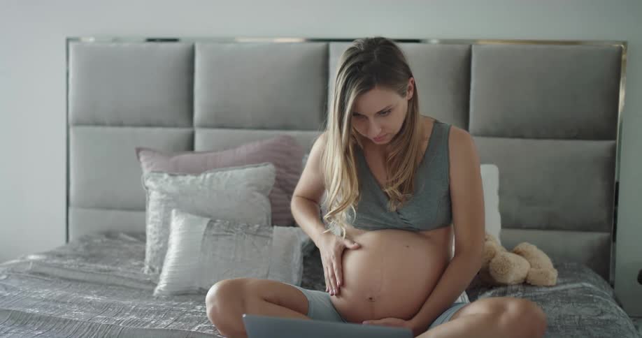 A pregnant woman rubs her stomach while using a laptop as she sits on a bed