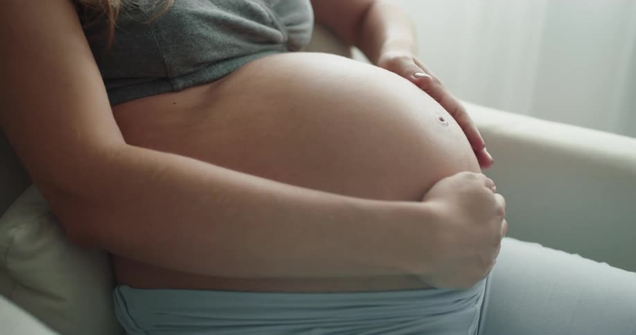 A pregnant woman rubs her stomach while sitting in an armchair indoors during the day