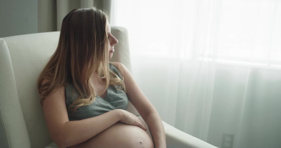 A pregnant woman rubs and cradles her stomach while sitting in a rocking chair indoors during the day
