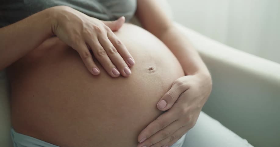 A pregnant woman rubs her stomach while sitting in an armchair indoors during the day
