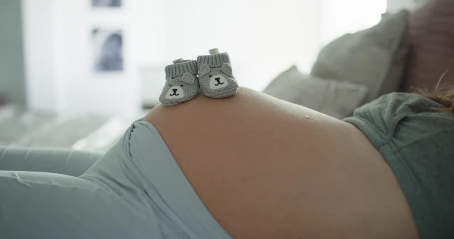 Baby boots resting over the belly of a pregnant woman resting in bed at home