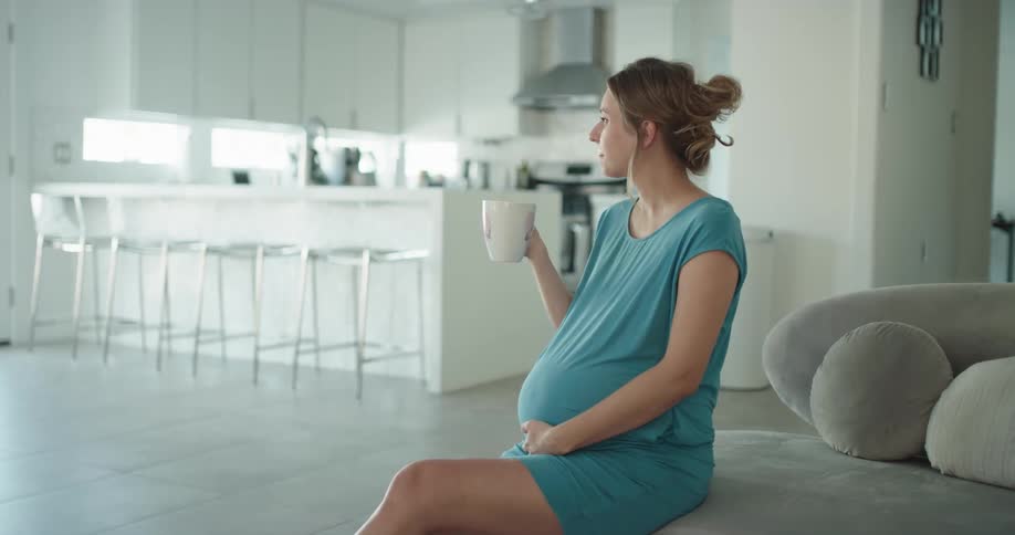 A pregnant woman is sitting on a couch and smiling while drinking from a mug