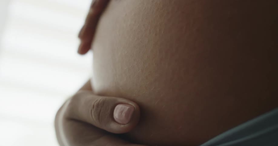 Pregnant woman strokes her belly next to window blinds