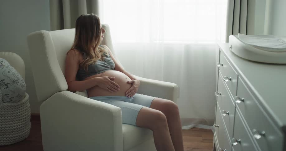 A pregnant woman strokes her belly while sitting in a rocking chair during the day