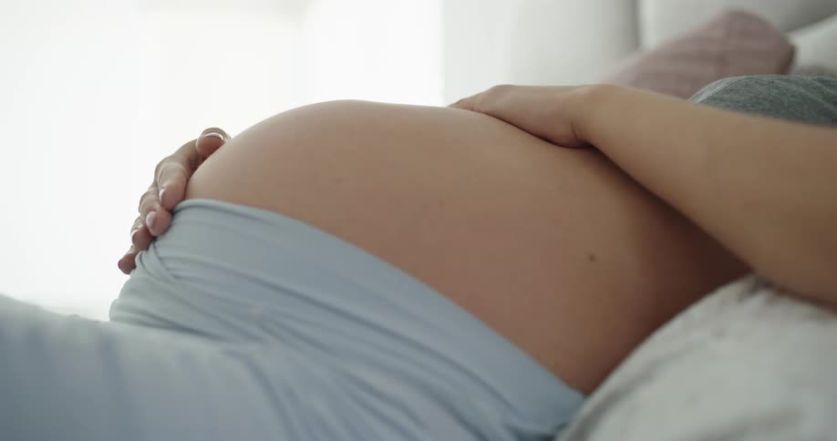 A pregnant woman strokes her stomach with her fingers while resting in bed during the day