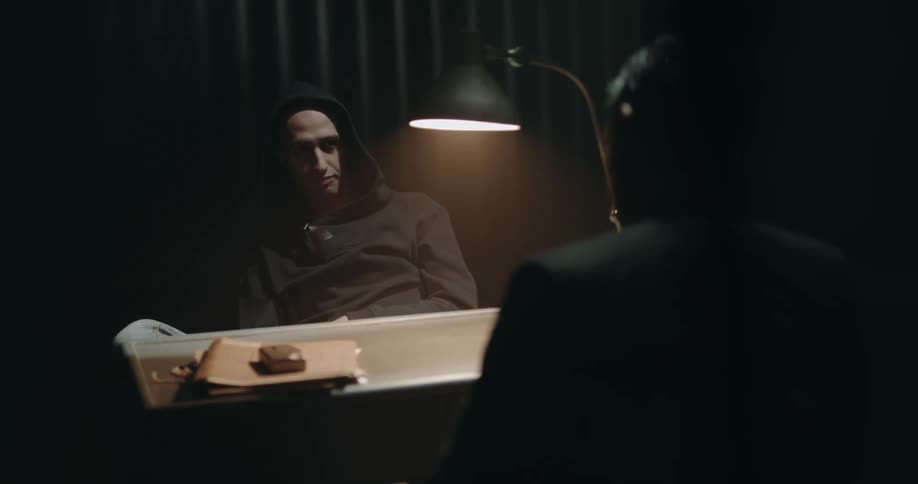 A man is sitting at a desk when another walks over and sits down and offers a cigarette