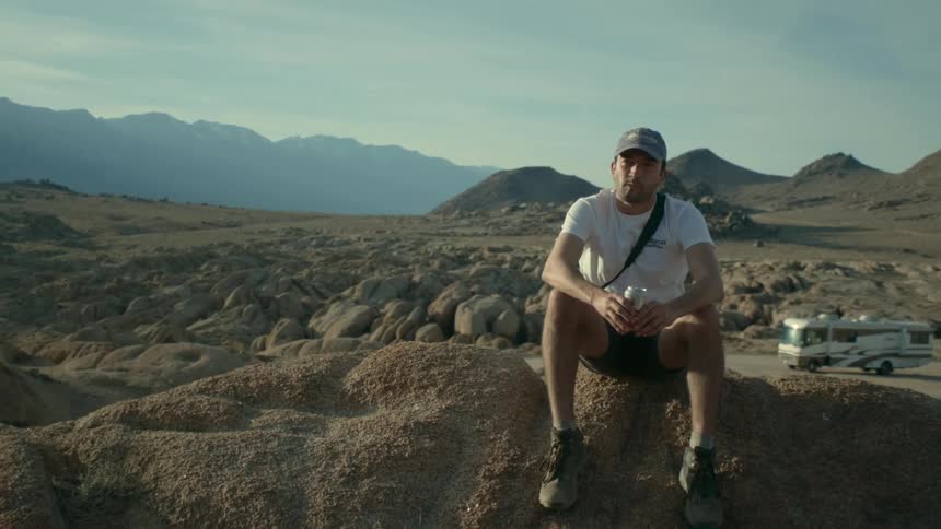 A man holds a can of beer while sitting on a rock formation in the desert during the day