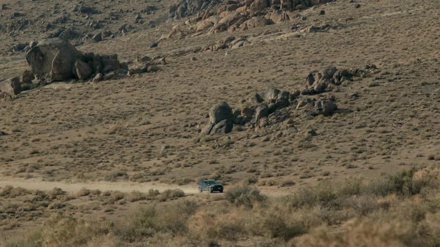 A vehicle travels along a dirt road through an arid countryside during the day