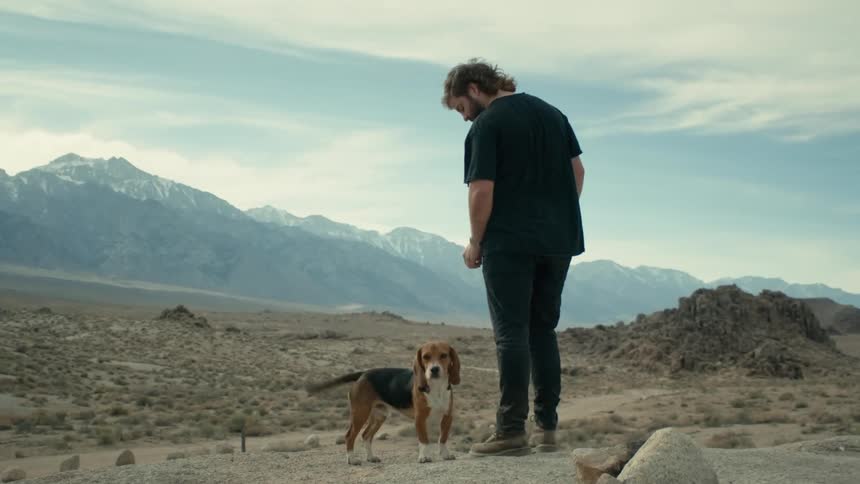 Man stands on a hill in the desert with his pet dog near the mountains